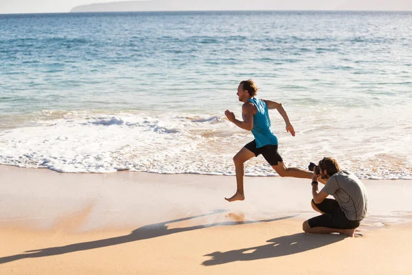 Behind the scenes of photo shoot of male sports athlete model running for photographer taking pictures for sport photoshoot. BTS on beach