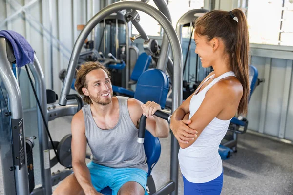 Fitness gym trainer talking to man training on workout equipment machine indoors. Couple happy working out together