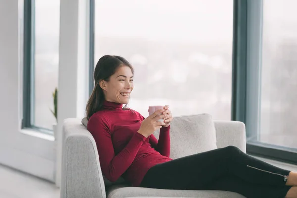 Ev yaşam tarzı Mutlu Asyalı kadın oturma odasında çay içerek rahatlıyor. Hafta sonları boş vakitlerin tadını çıkarıyor. Pencere manzarasında kanepede oturuyor. İç konfor — Stok fotoğraf
