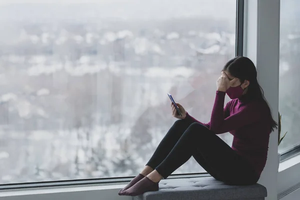 Telefoon gebruiken trieste vrouw dragen masker alleen thuis tijdens het coronavirus. Depressie, verslaving aan sociale media, angst, depressie, verdriet mentale warmte concepten — Stockfoto