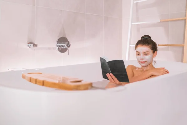 Bath at home Asian woman pampering relaxing taking a hot bath to relax and destress. Reading a book and putting facial face mask treatment happy zen lifestyle — стоковое фото