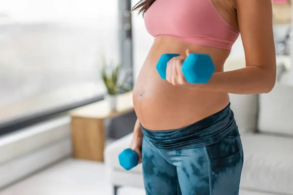 Pregnant woman fitness strength training exercise with dumbbells at home. Body weights workout arm curls closeup of belly