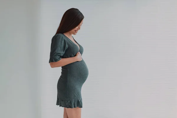 Schöne schwangere junge Asiatin, die in grünem Kleid auf ihren Bauch zum Umstandsmode-Fotoshooting herabblickt. Schwangerschaftsfotos im dritten Trimester — Stockfoto