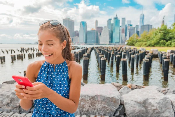 New York tourist woman using app on phone by Manhattan city skyline waterfront. People walking enjoying view of downtown from the Brooklyn Bridge park Pier 1 salt marsh — стокове фото