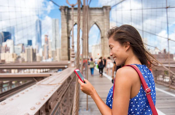 纽约的女游客在布鲁克林桥上使用电话应用程序向曼哈顿天际线走去。年轻的多文化女性专业人员，美国纽约市 — 图库照片