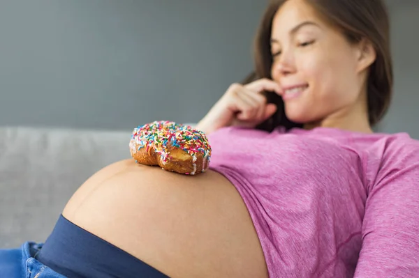 Unhealthy food during pregnancy. Sugar temptation craving Asian pregnant woman trying to resist eating a sweet donut on her baby belly. Funny eating cravings concept.