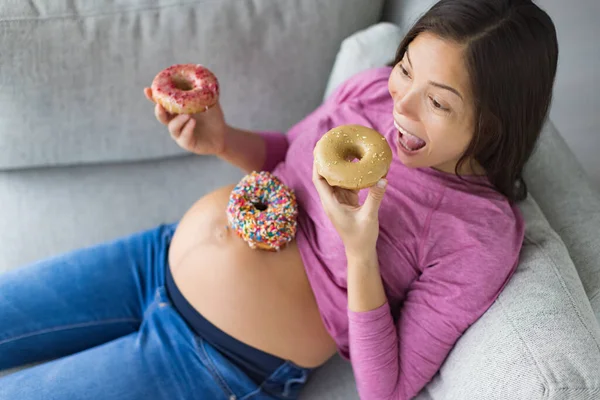 Pregnancy food cravings funny Asian pregnant woman eating donuts at home hungry for dessert cakes and sweet snacks. Gestational diabetes, nutrition, weight gain.