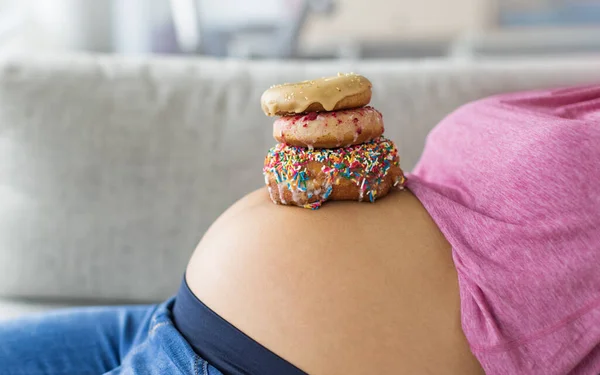 Pregnant woman with donuts on belly. Cravings of desserts and sweets during pregnancy, Girl eating unhealthy pastries on baby bump for gestational diabetes.