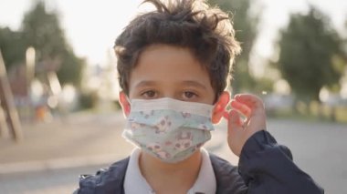 Sevimli mutlu çocuk kameraya bakıyor. Bebek etrafına bakıyor, sonra gülümsüyor ve gülüyor. 5 yaşında, açık renk saçlı, kahverengi gözlü bir çocuk parkta gülümsüyor. Ağır çekim. Maskesini çıkarıyor..