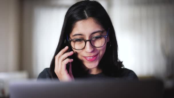 眼鏡をかけたかなり若いブルネットの女の子は 現代的な部屋のソファに座って コンピュータを見ている間に電話で話しています オンライン作業 — ストック動画