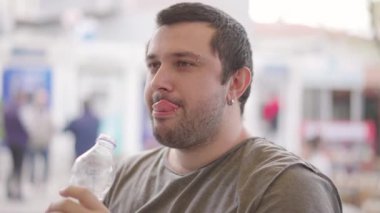 Şehrin arka planında evcil hayvan şişesinden su içen genç adam gülümsüyor ve iyi hissediyor. Eğlence ve gülümseme. tatlı su içmek