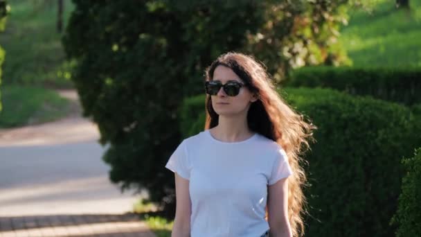 Молодая красивая женщина в белой футболке и солнечных очках гуляет по парку летом — стоковое видео