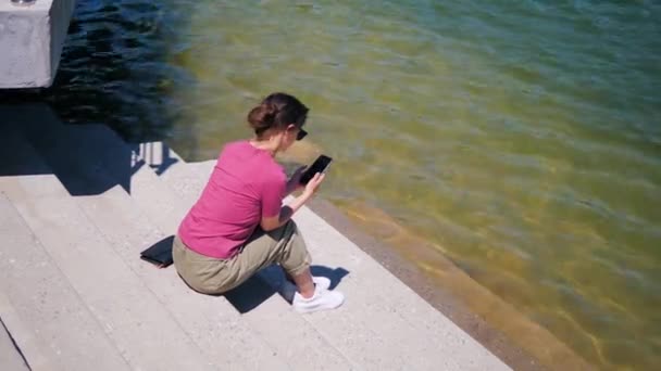 湖のそばに座って晴れた夏の日に携帯電話を使う若い女性は — ストック動画