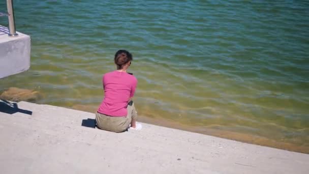 Mujer joven sentada en los escalones junto al lago y usando un teléfono móvil en un día soleado de verano — Vídeo de stock
