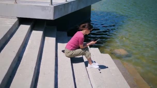 Jovem mulher sentada nos degraus junto ao lago e usando um telefone celular em um dia ensolarado de verão — Vídeo de Stock