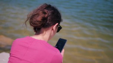 Gölün kenarındaki merdivenlerde oturan ve güneşli bir yaz gününde cep telefonu kullanan genç bir kadının arka görüntüsü.