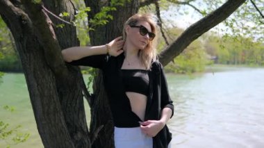 Siyah bluzlu, güneş gözlüklü ve düğmesiz gömlekli güzel bir kadın ağacın altında duran gölün yanında poz veriyor.