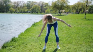 Genç bayan sporcu, güneşli bir günde, yazın göl kenarındaki yeşil çimlerde ısınma egzersizleri yapıyor..