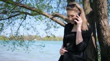 Güzel sarışın bir kadın, caddede, bir gölün kenarında telefonla konuşuyor ve telefonu kapatıyor.