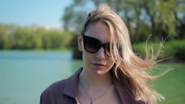 Portrait de femme blonde en lunettes de soleil sur le fond du lac dans le parc d'été. Les cheveux se développent dans le vent. — Video