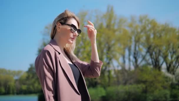 Jeune belle femme en lunettes de soleil marche à travers le parc en été et regarde l'heure sur l'horloge — Video