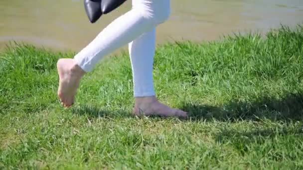 The feet of a young girl barefoot walking on the green grass in summer on a sunny day along the lake. Side view — ストック動画