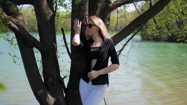 Une belle jeune femme blonde en lunettes de soleil noires en vêtements d'été pose par un arbre sur le fond d'un lac, la nature — Video