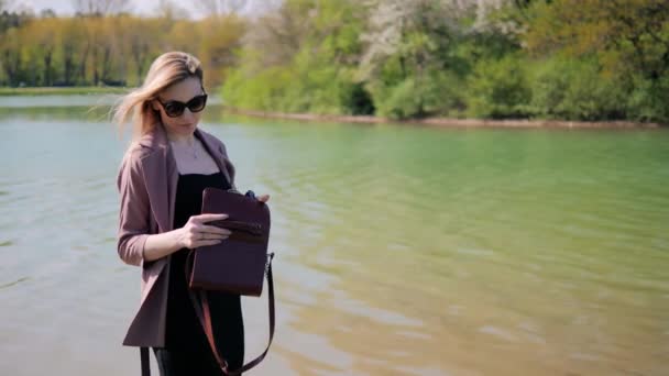 Ung vacker kvinna lägger något i sin handväska mot bakgrund av en sjö i en stadspark på våren — Stockvideo