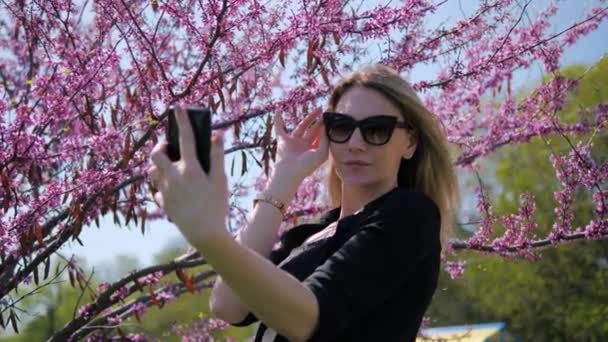 A pretty young blonde woman in sunglasses takes a selfie on her phone on a beautiful background of cherry blossoms — Vídeo de Stock
