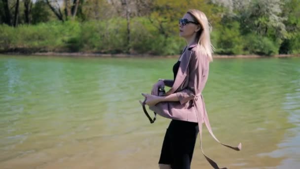 Young beautiful woman hangs a handbag on her shoulder while walking along the lake in the park — Stockvideo