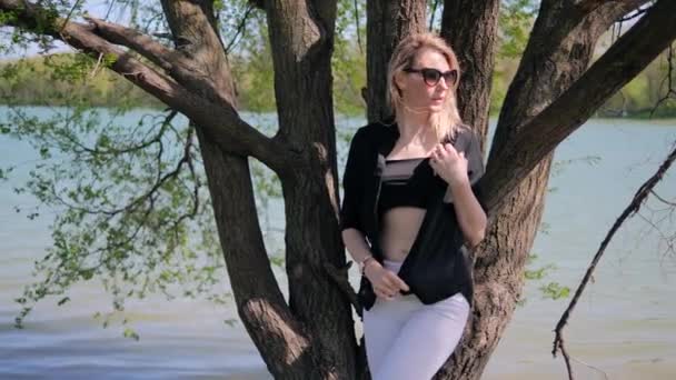 A blonde woman in sunglasses in a black top and shirt in white jeans stands by a tree by the lake in the park in summer — ストック動画