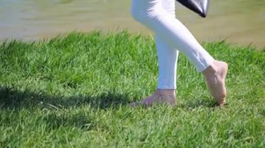 Yeşil çimenlerde yürüyen ve elinde ayakkabıları olan genç bir kadının çıplak ayaklarının yakın çekimi.