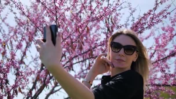 A young beautiful woman takes a selfie admiring herself against the background of a blooming pink Sakura tree — 图库视频影像