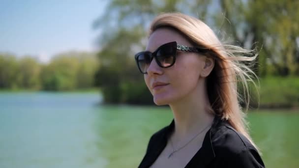 Retrato de cerca de una joven rubia con gafas de sol negras caminando en el parque sobre el fondo del lago y los árboles en un día de verano — Vídeos de Stock