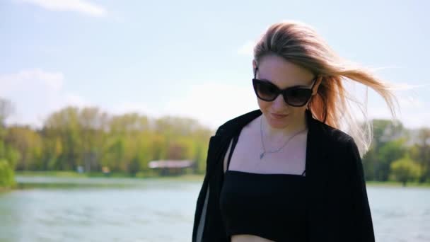 Portrait of a young blonde woman in black sunglasses walking in the park along the lake on a hot summer day — 图库视频影像