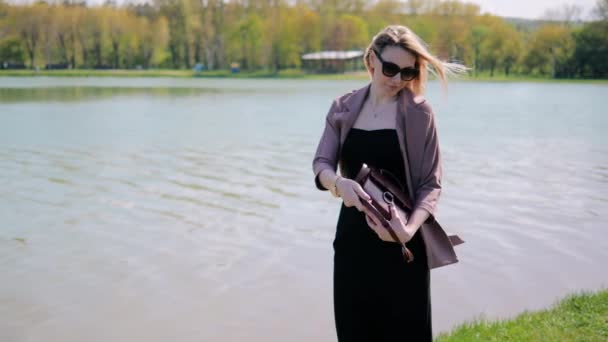Une belle jeune femme dans un parc au bord du lac est à la recherche d'un miroir dans son sac à main pour devenir plus jolie — Video