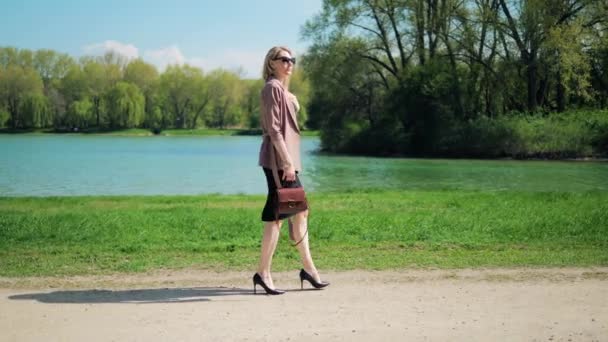 Young businesswoman with a handbag walks along the lake through the park and looks at the watch in her arms. — ストック動画