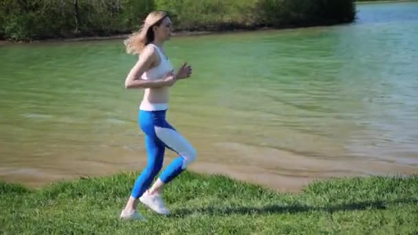 Sportowa kobieta biega na świeżym powietrzu wzdłuż jeziora w parku latem w słoneczny dzień — Wideo stockowe