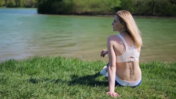 Joven atleta femenina está sentado junto a la orilla del lago en la hierba en el parque en verano en el día soleado — Vídeo de stock