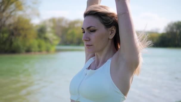 A fiatal női sportoló felmelegíti a kezét a friss levegőn. Gyakorlatok a parkban a tó mellett nyáron a napsütéses napon — Stock videók