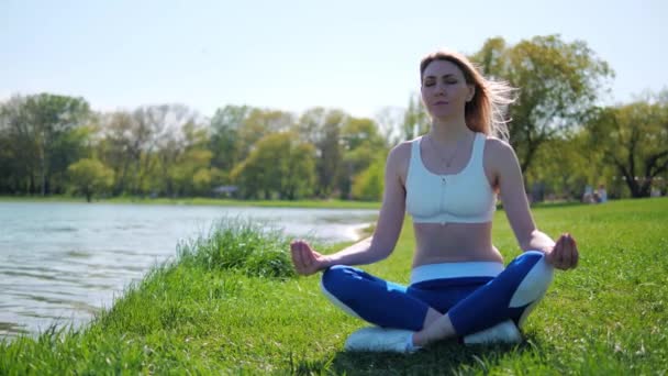 Молодая блондинка в спортивной одежде сидит в позе лотоса и медитирует на открытом воздухе в парке у озера летом в солнечный день. — стоковое видео