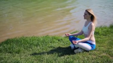 Dişi bir atlet, göl kıyısında çimlerin üzerinde lotus pozisyonunda oturuyor. Kız dışarıda parkta yoga yapıyor..