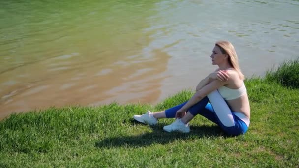 Young female athlete is sitting in park by lake shore on grass and resting after sports workout — Stock Video