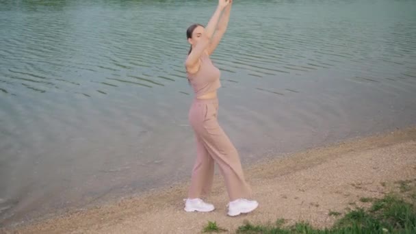 Hermosa joven en ropa deportiva está bailando posando a lo largo de la orilla del lago en el parque — Vídeos de Stock