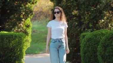 Güneş gözlüklü güzel bir esmer kadın yeşil ağaçların arasında bir yaz parkında yürüyor..