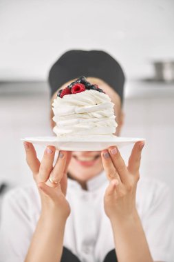 Üniformalı şekerci elinde bir pavlova tatlısı tutuyor, mutfakta gösteriyor. dikey çerçeve, metin için alanı kopyala