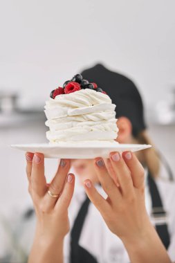 Üniformalı şekerci elinde bir pavlova tatlısı tutuyor, mutfakta gösteriyor. dikey çerçeve, metin için alanı kopyala