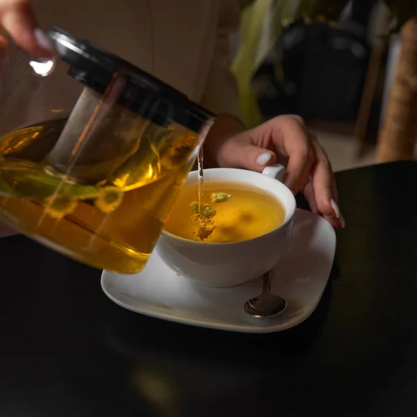 やかんからお茶をゆっくりと磁器のカップに注ぎます 朝の朝食時に台所で緑茶の準備をしている若い女性は ティーポット ティーカップ 健康なハーブの葉を使用して — ストック写真