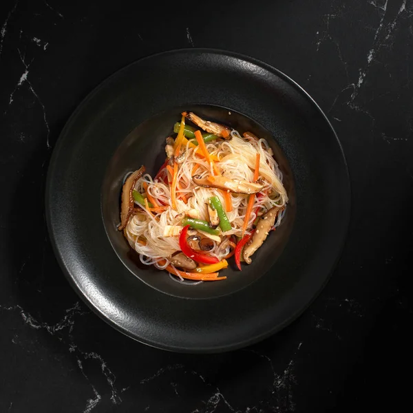 Manzaralı, sebzeli Funchoza. Sebzeli lezzetli şehriyeler, koyu taşlı arka planda. Geleneksel Asya yemekleri, kavrulmuş erişte ya da sıcak buharlı siyah bir kasede rafadan erişte.