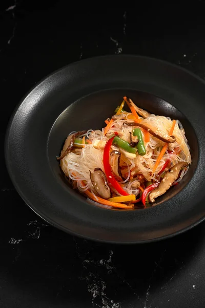 Manzaralı, sebzeli Funchoza. Sebzeli lezzetli şehriyeler, koyu taşlı arka planda. Geleneksel Asya yemekleri, kavrulmuş erişte ya da sıcak buharlı siyah bir kasede rafadan erişte.